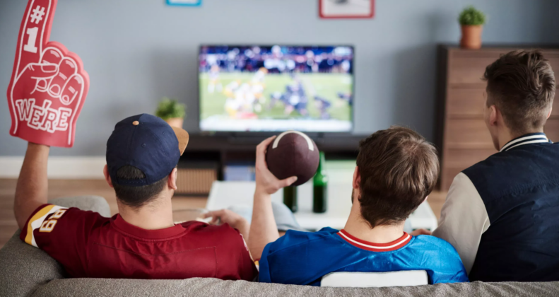 Three guys sitting on a couch watching football, holding a foam finger and a football